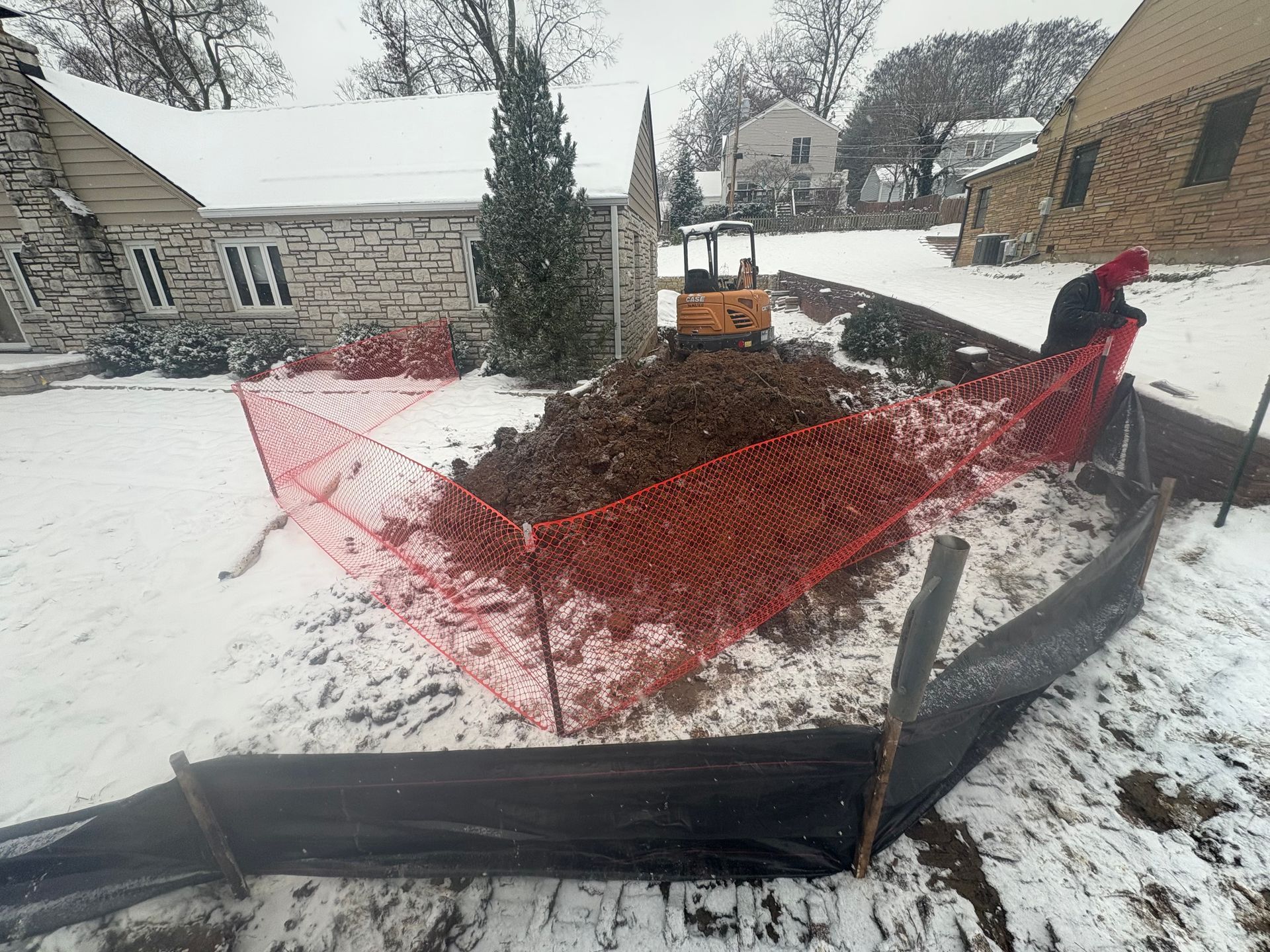 Snowy backyard construction site with a mound of dirt, orange machine, and red safety fencing.