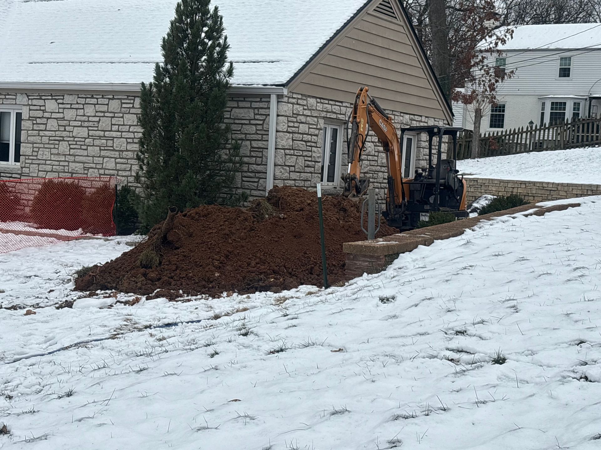 Backhoe digging a trench beside a house in a snowy yard, with a large dirt pile nearby.