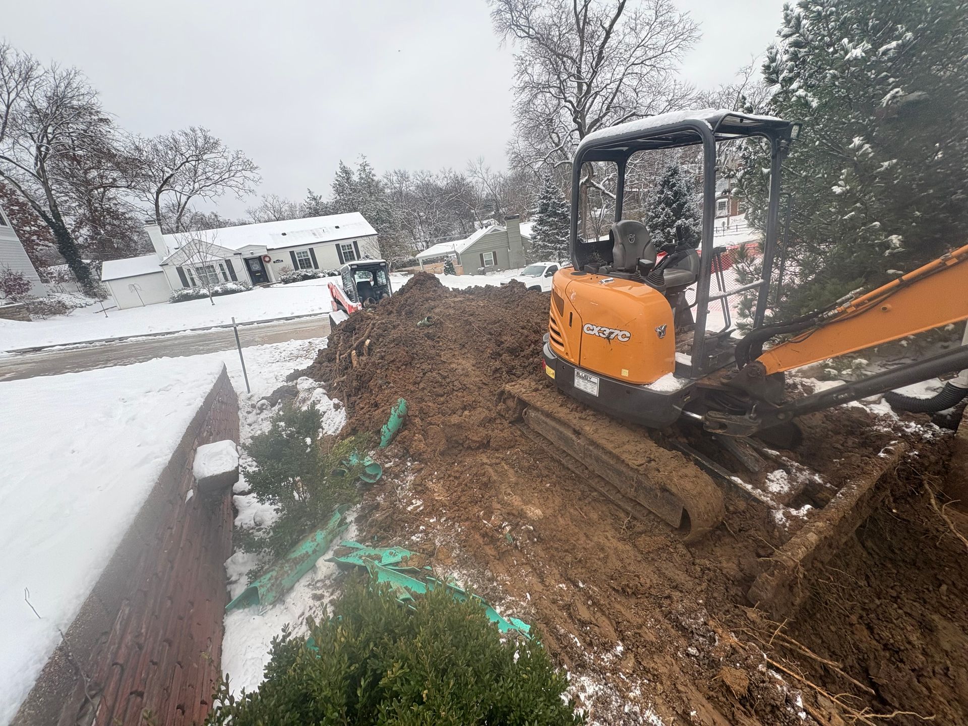 Orange excavator digging mud beside a snowy road in a residential area.