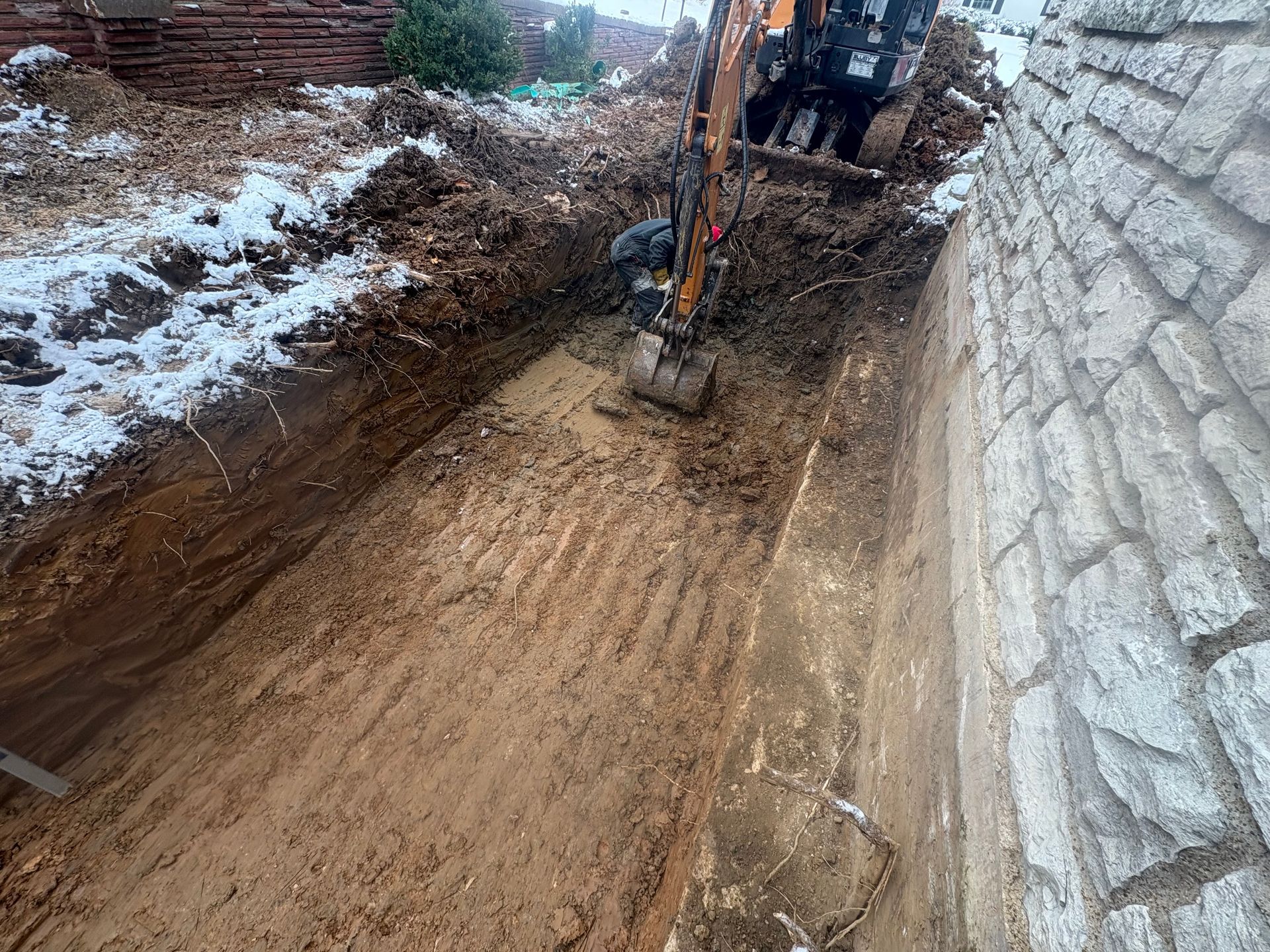 Excavator digging a muddy trench beside a stone wall with snow on the ground.