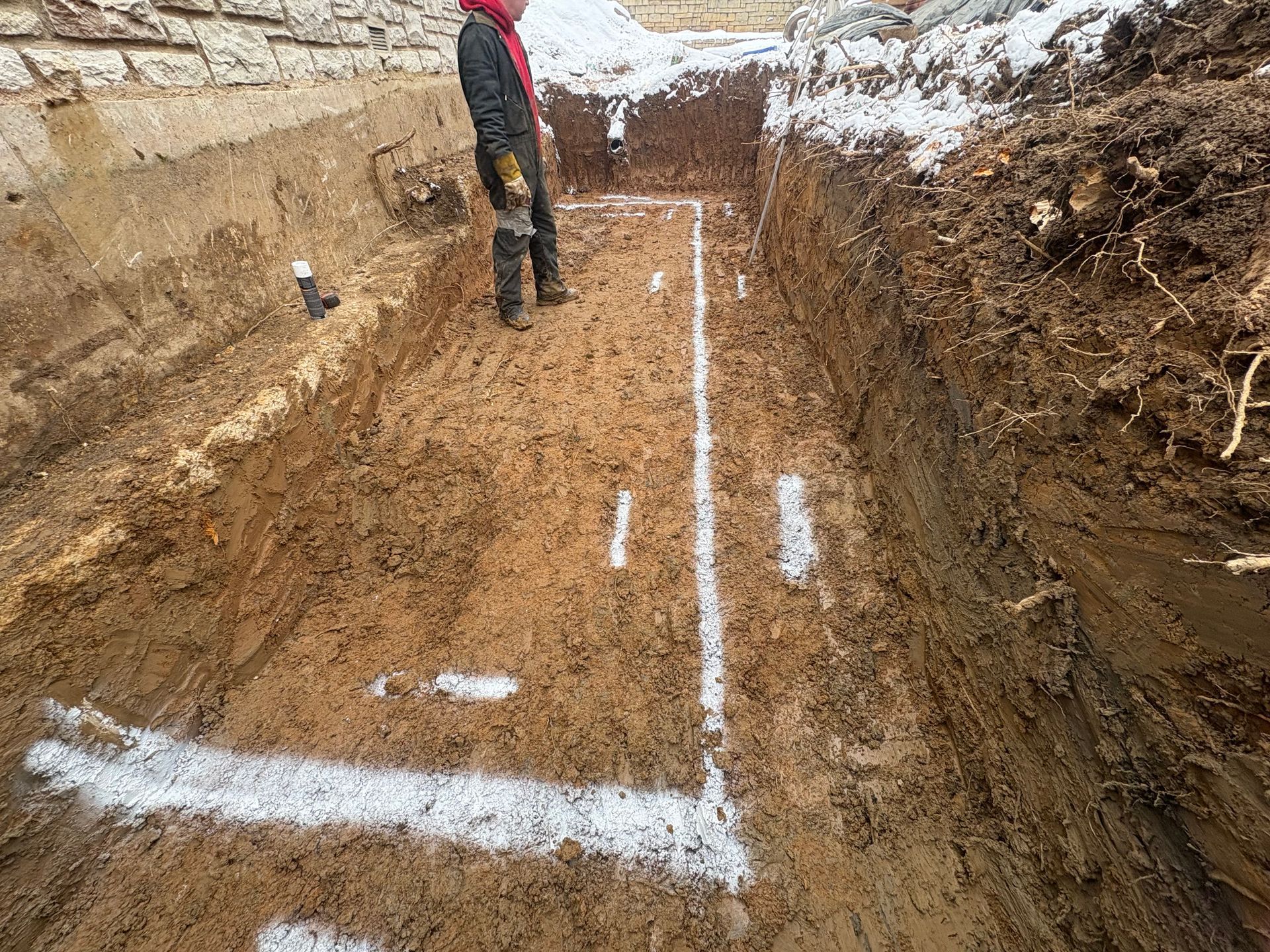 Trench excavation with white layout lines and a worker standing at the edge in winter soil.