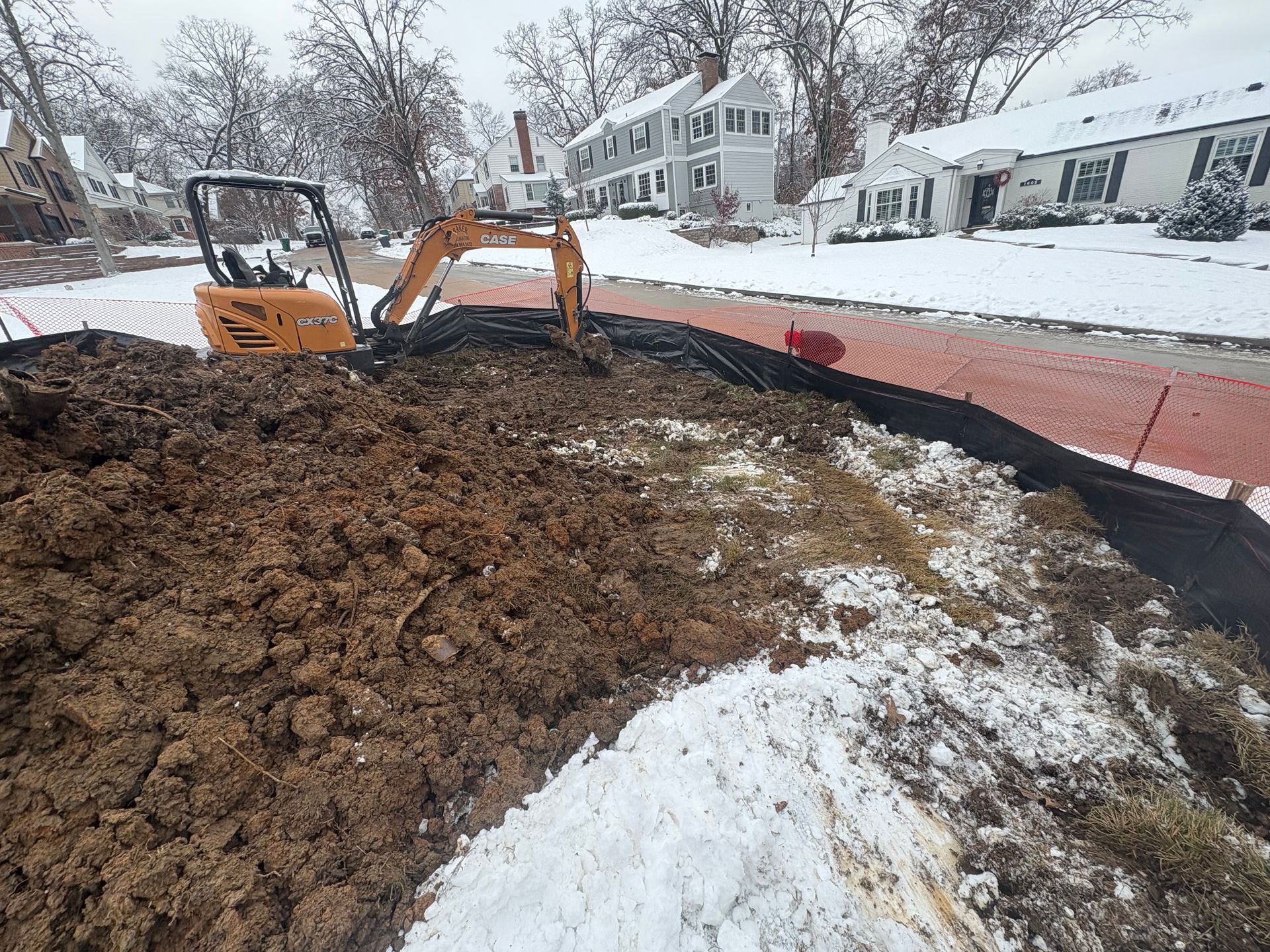Excavator on muddy lot beside snow-covered street and houses, with orange safety fence in front.