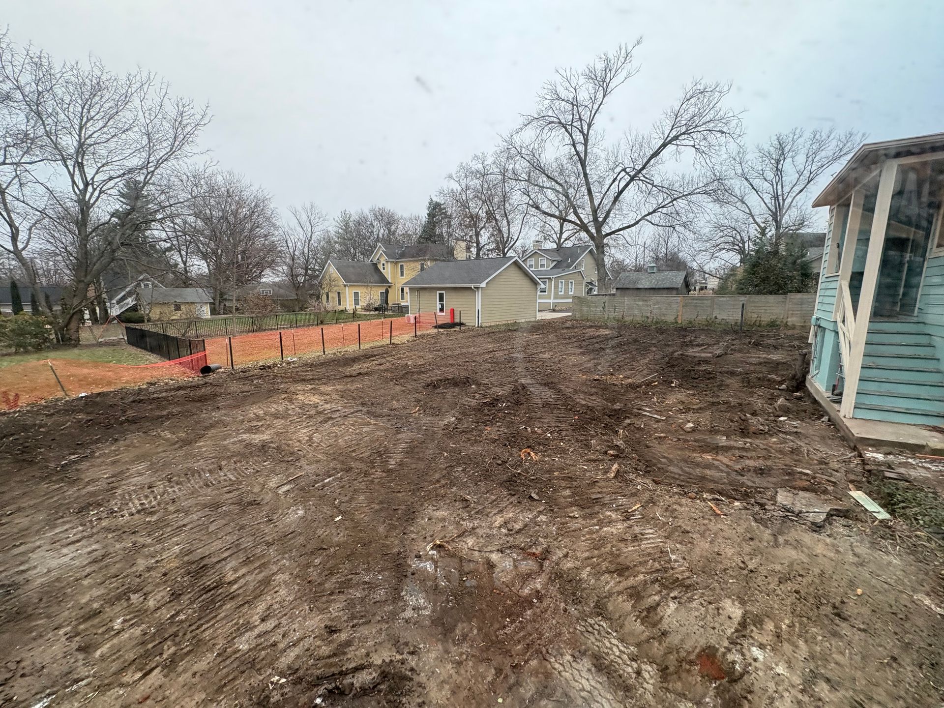 Dirt-covered backyard with leafless trees, orange fencing, and a small house in the distance.