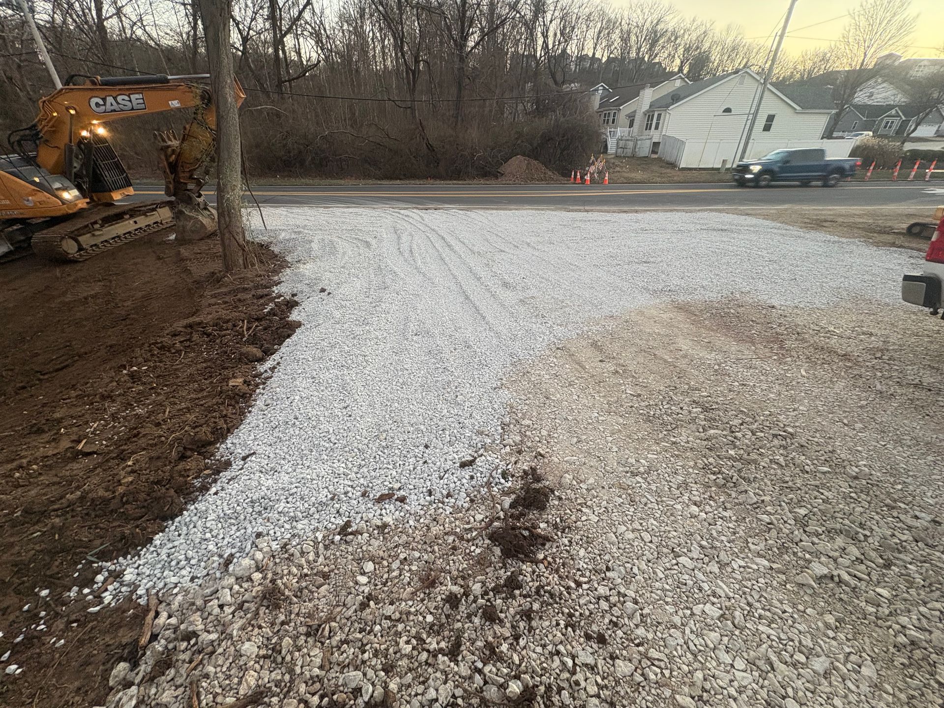 Gravel driveway under construction beside excavator, with dirt pile and houses in the background.