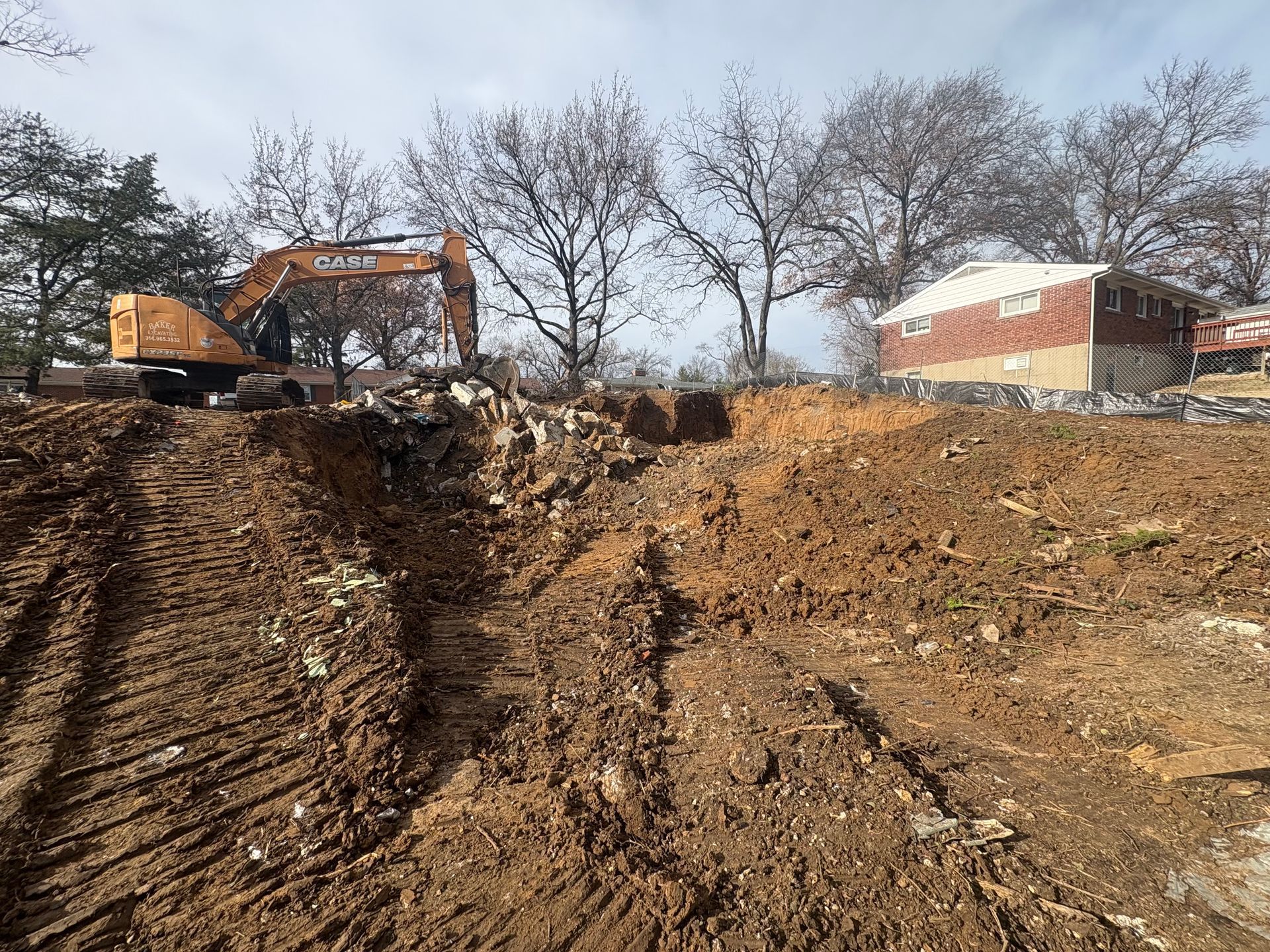 Excavator on a dirt embankment at a construction site with bare trees and a brick building nearby.