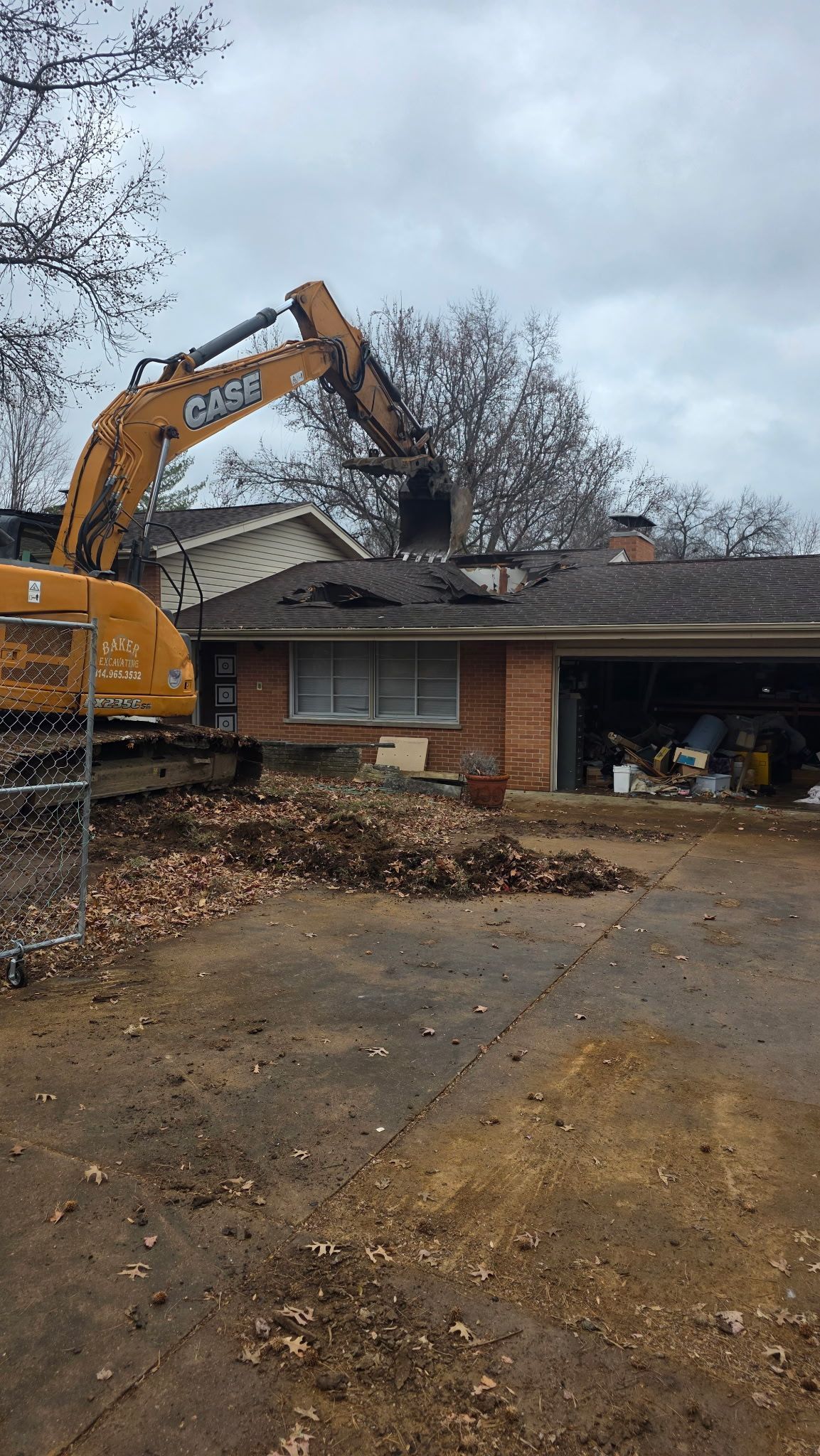 Yellow excavator demolishing a roof on a house in a muddy residential driveway.