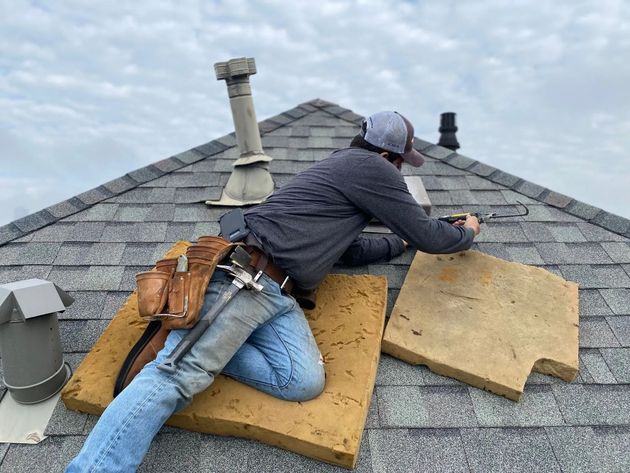 Roofing worker kneeling on a shingled roof, installing plywood panels near a chimney.