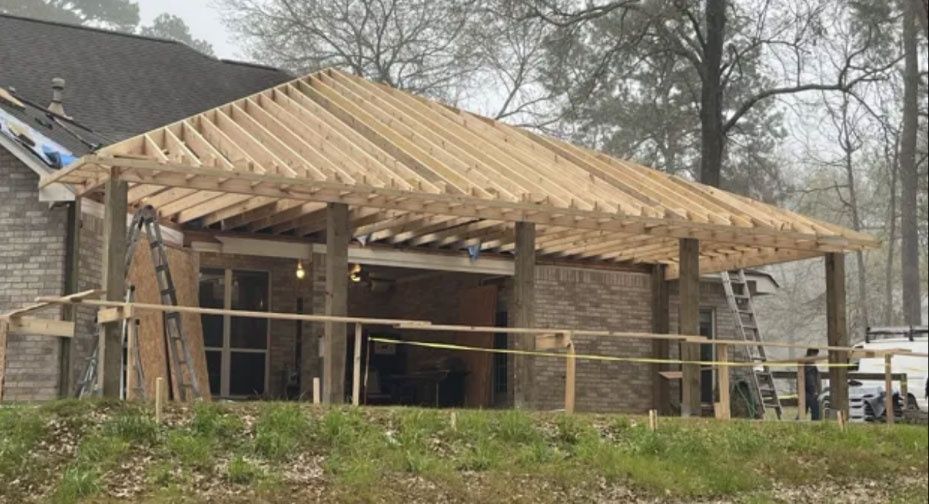 Partially built house with exposed wooden roof framing and open front porch in a wooded area