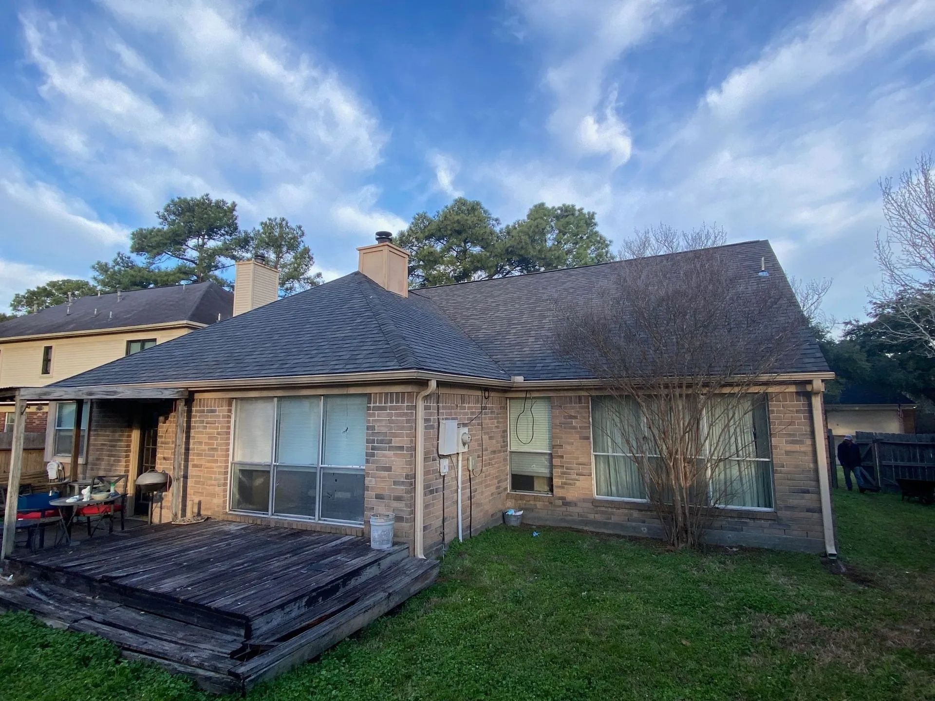 Backyard view of a brick house with a dark roof, patio deck, and cloudy sky