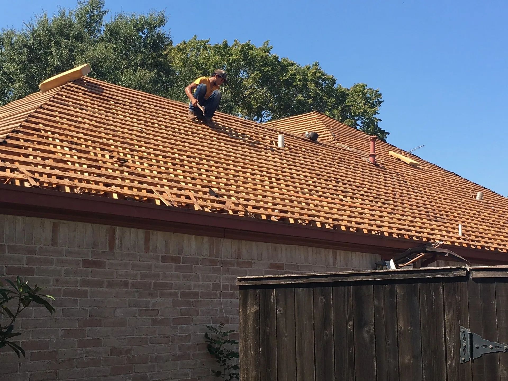 Worker on a terracotta-tiled roof above a brick building, with trees and blue sky in the background.