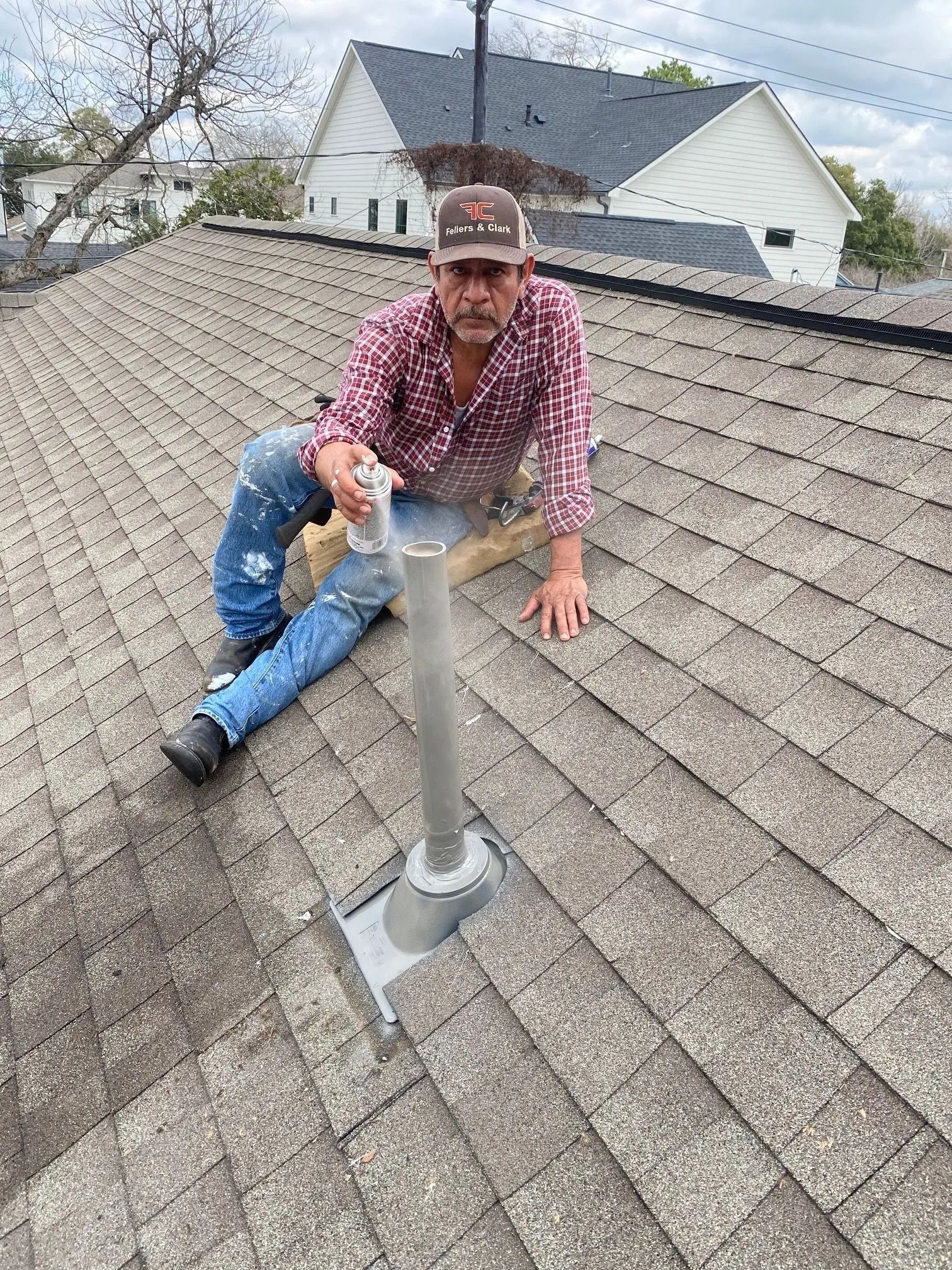Man kneeling on a shingled roof beside a vent pipe, repairing the flashing.