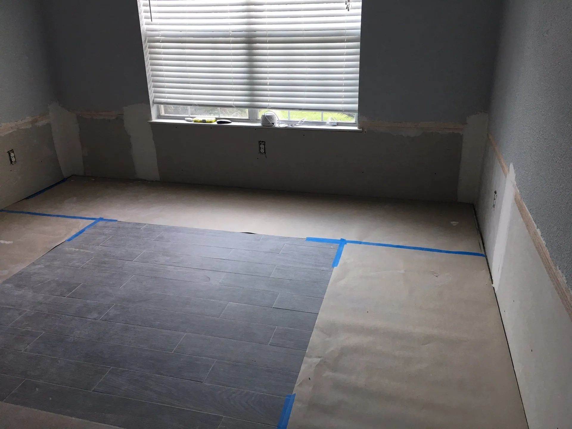 Empty room with gray walls, window blinds, and taped-off flooring sections under renovation