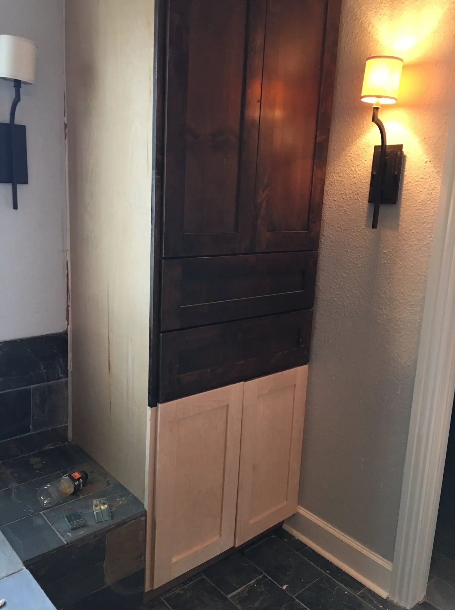 Tall dark wood wall cabinet with light wood doors in a hallway, beside wall sconces and a tiled step.
