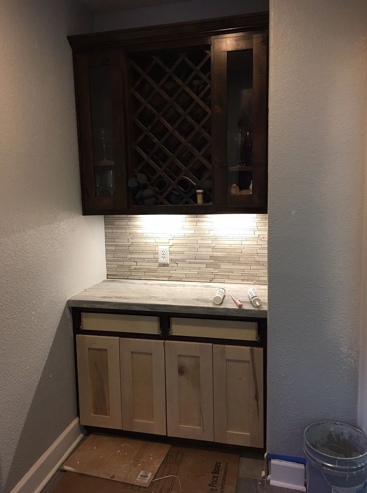 Small hallway wet bar with dark cabinets, stone backsplash, sink, and unfinished lower cabinets.