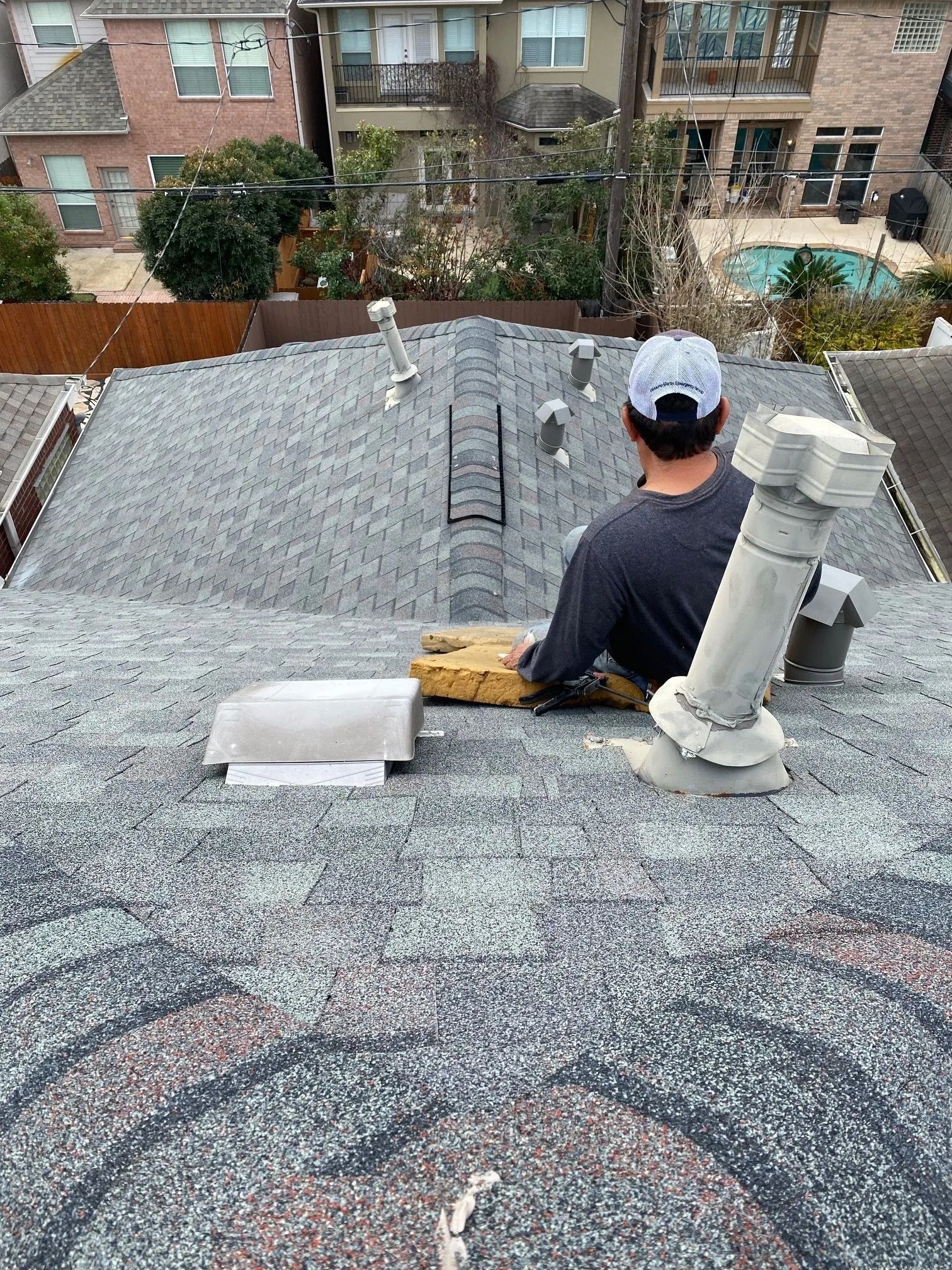 Worker on a gray shingle roof near a chimney, overlooking a backyard with neighboring homes.