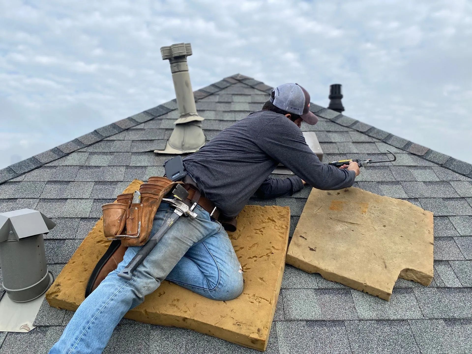 Worker repairing roof shingles while lying on a steep gray shingled roof