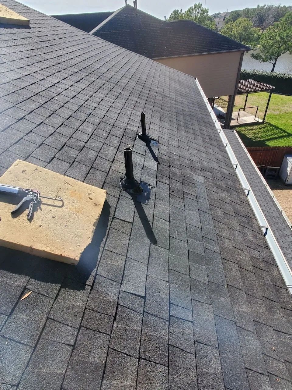 Roof with damaged shingles and a loose vent pipe on a sloped house roof, near a yard.