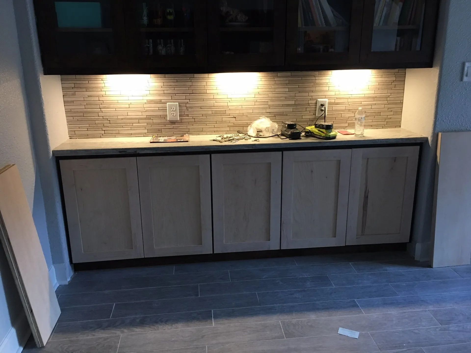 Built-in kitchen counter with under-cabinet lights and gray wood cabinets against a stone backsplash.
