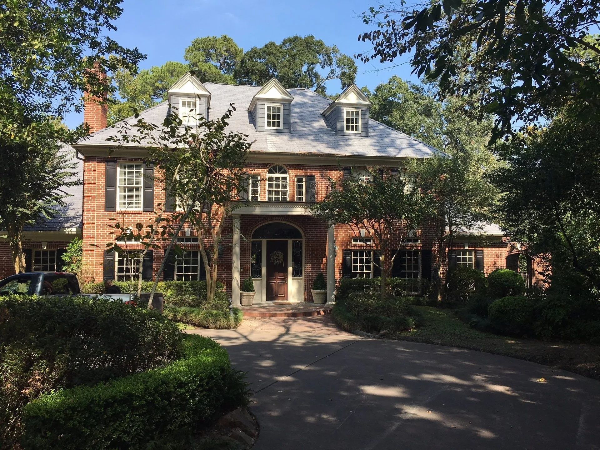 Large brick colonial house with white trim, dormer windows, and a tree-lined driveway in front