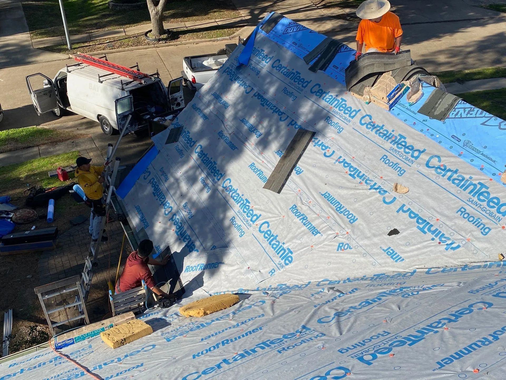 Workers installing roofing underlayment on a steep house roof, with tools and a truck nearby.