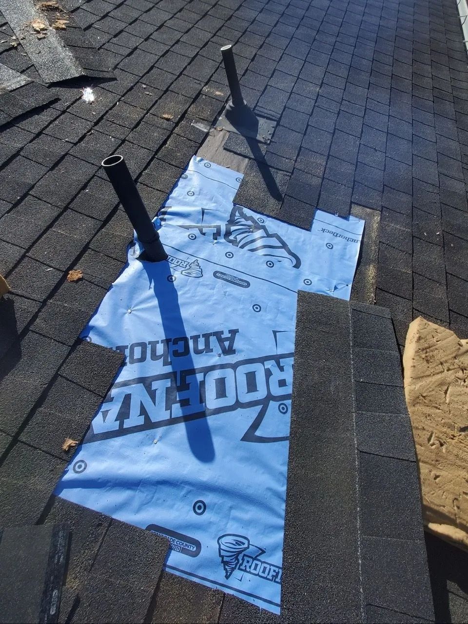 Blue roofing underlayment torn on a shingled roof, with black vent pipes nearby.