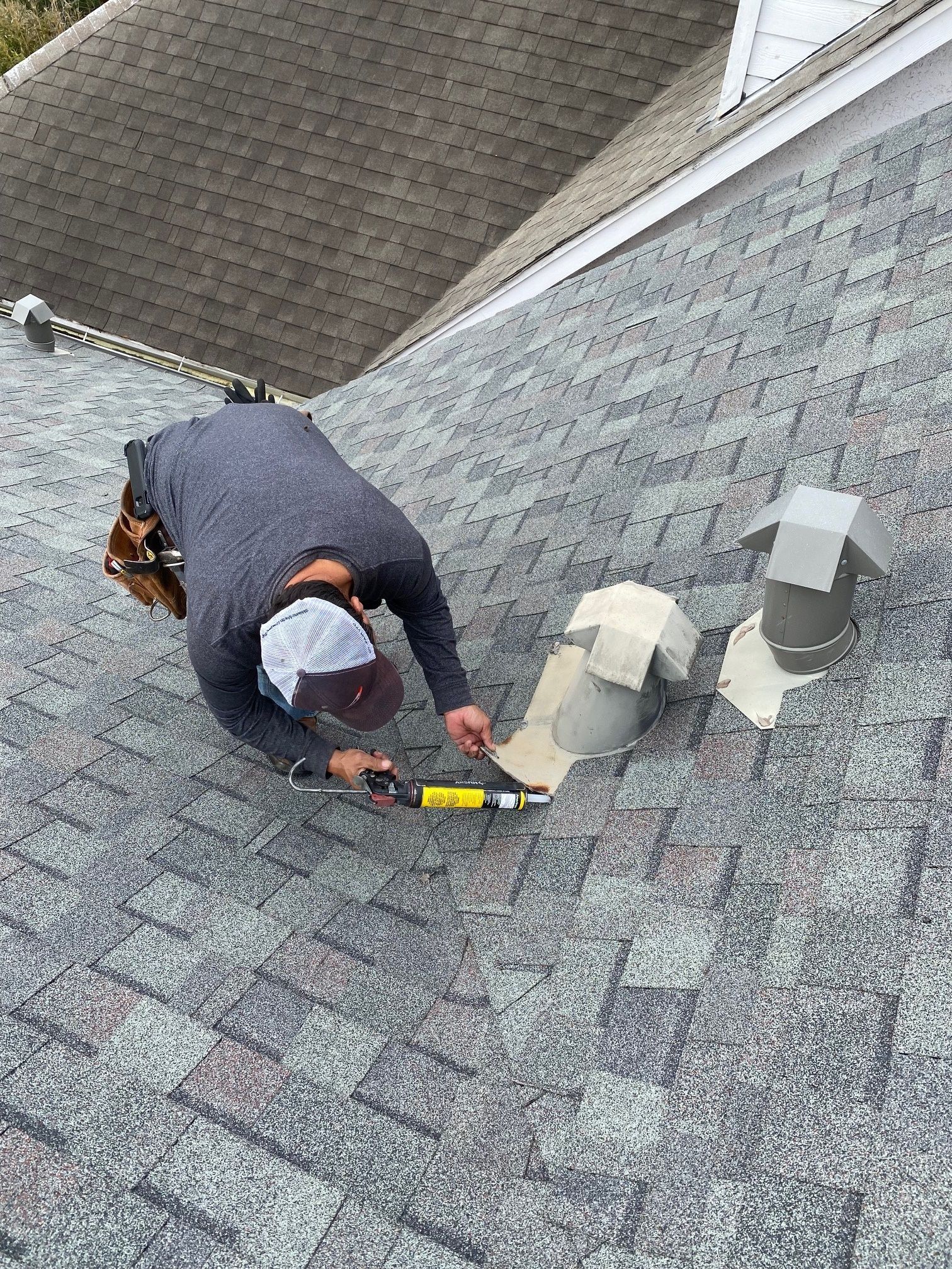 Worker repairing a gray shingle roof near rooftop vents with yellow caulking tool