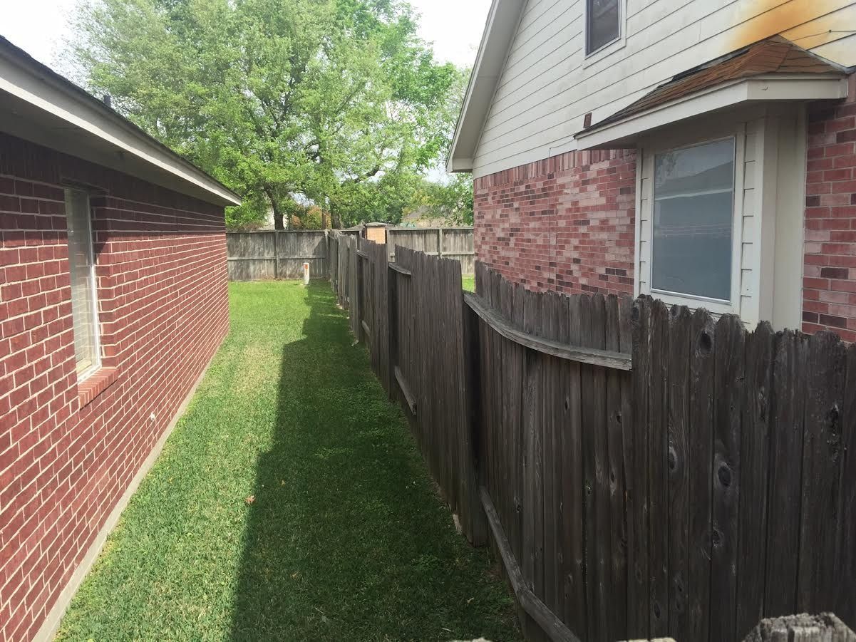 Narrow grassy side yard between a brick house and a wooden fence, with neighboring house in view.