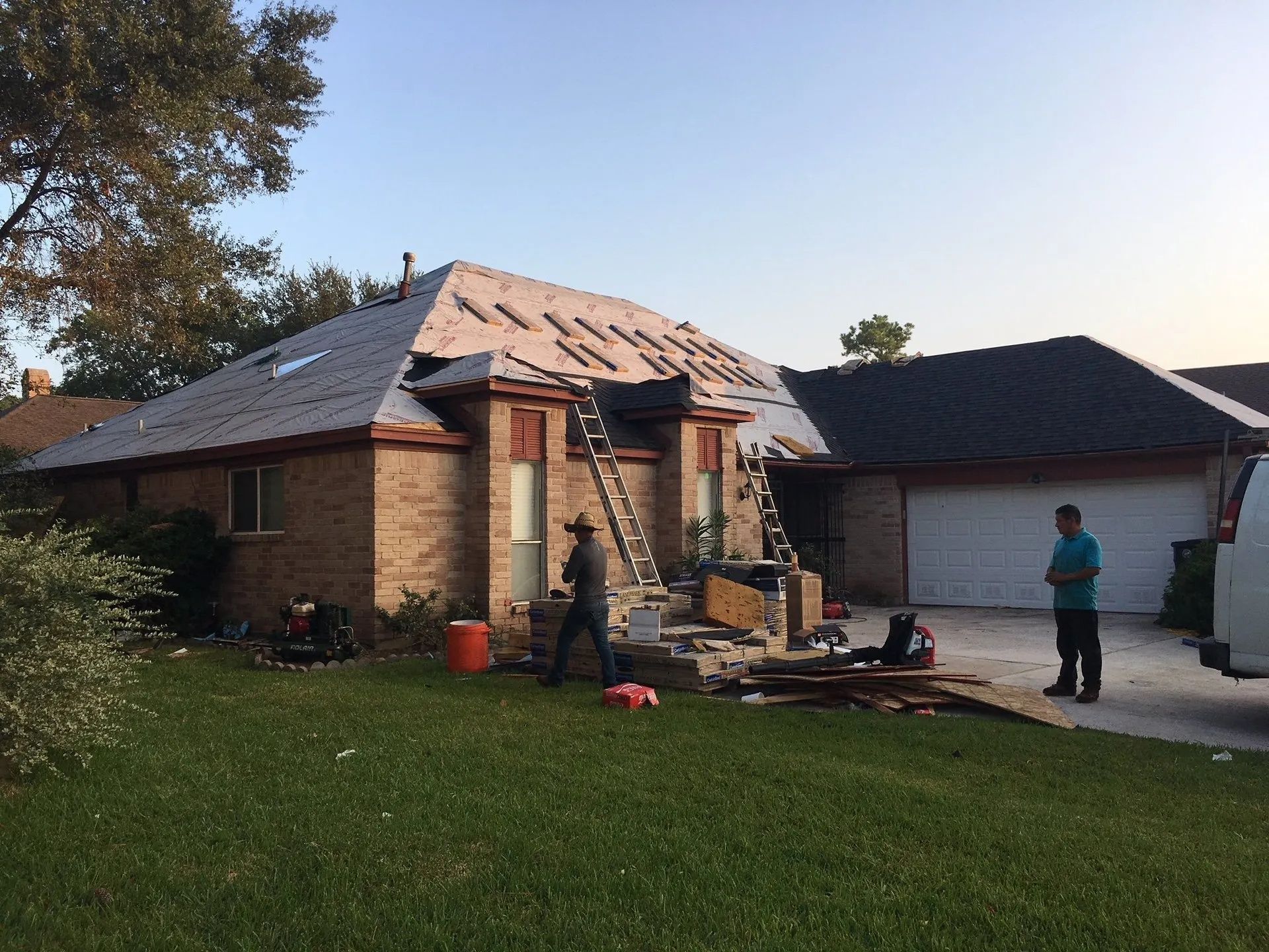 Damaged brick house with missing roof tiles; two people stand near the driveway at dusk.