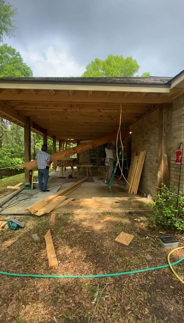 Construction site under a covered carport with workers, lumber, and tools beside a stone house