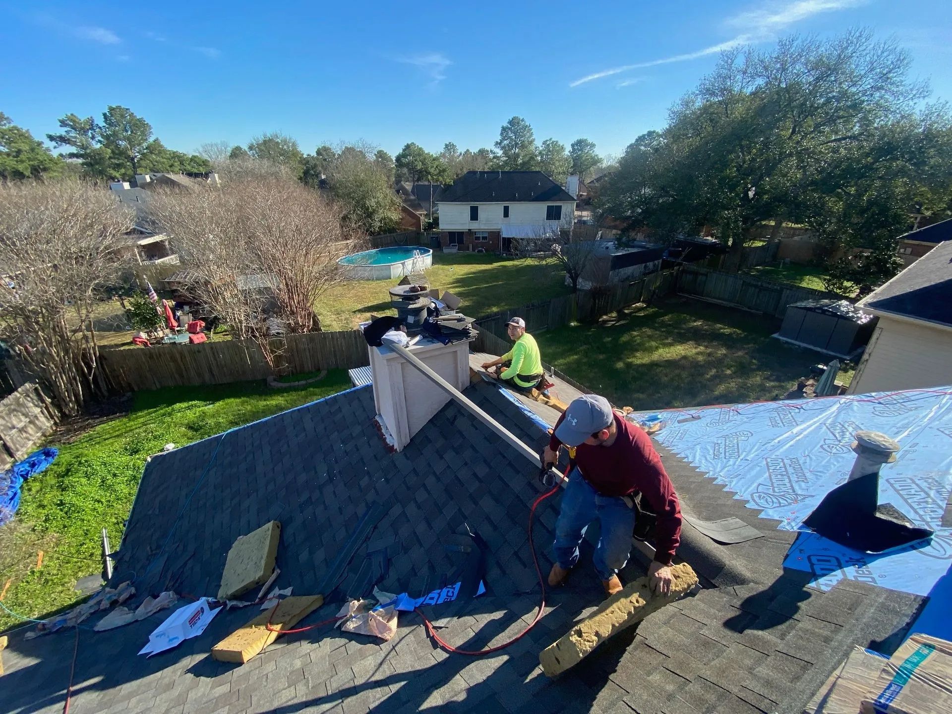 Workers repairing a roof on a house, with shingles, tools, and a backyard in the background