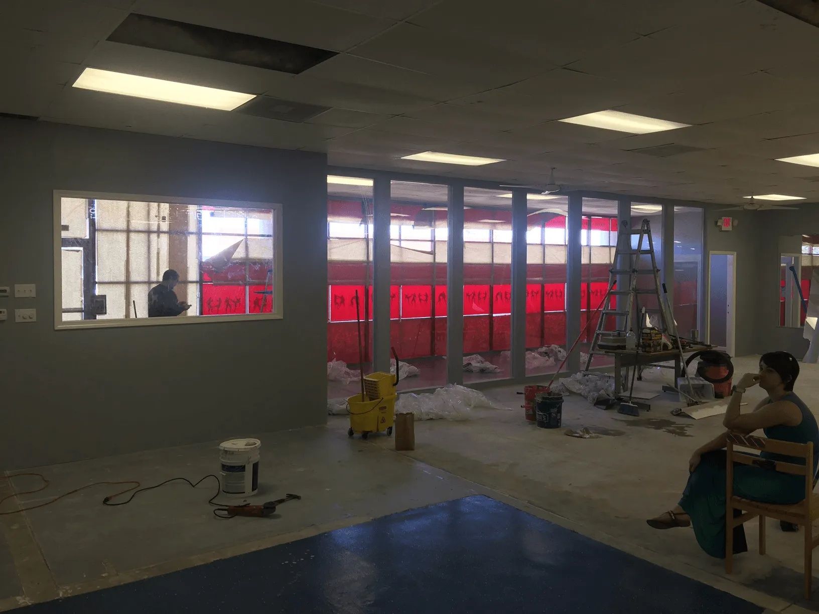 Empty office under renovation with ceiling lights, glass walls, scattered tools, and one person seated at right