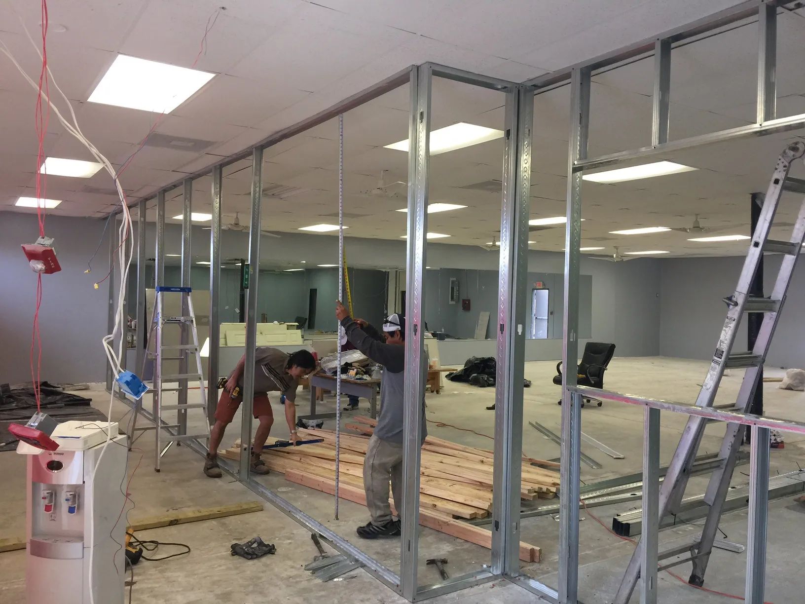 Workers installing glass office partitions inside a room under renovation, with ladders and tools visible.