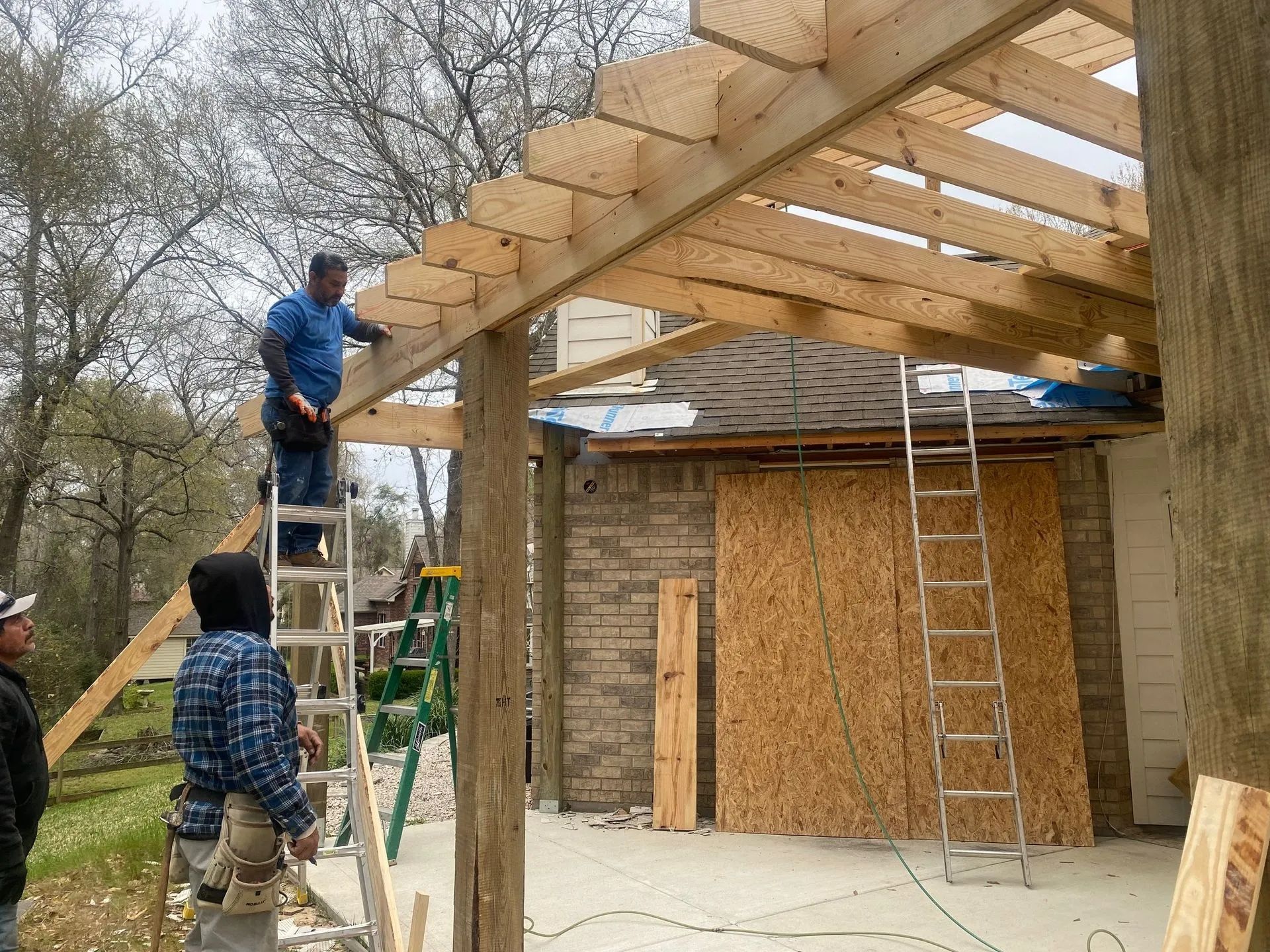 Workers building a wooden roof frame over a small structure outdoors, with ladders and plywood walls.