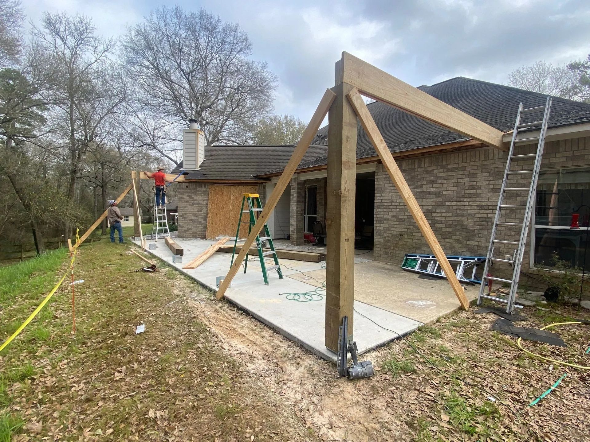 Workers building a wooden patio frame beside a stone house, with ladders and tools on a concrete slab.
