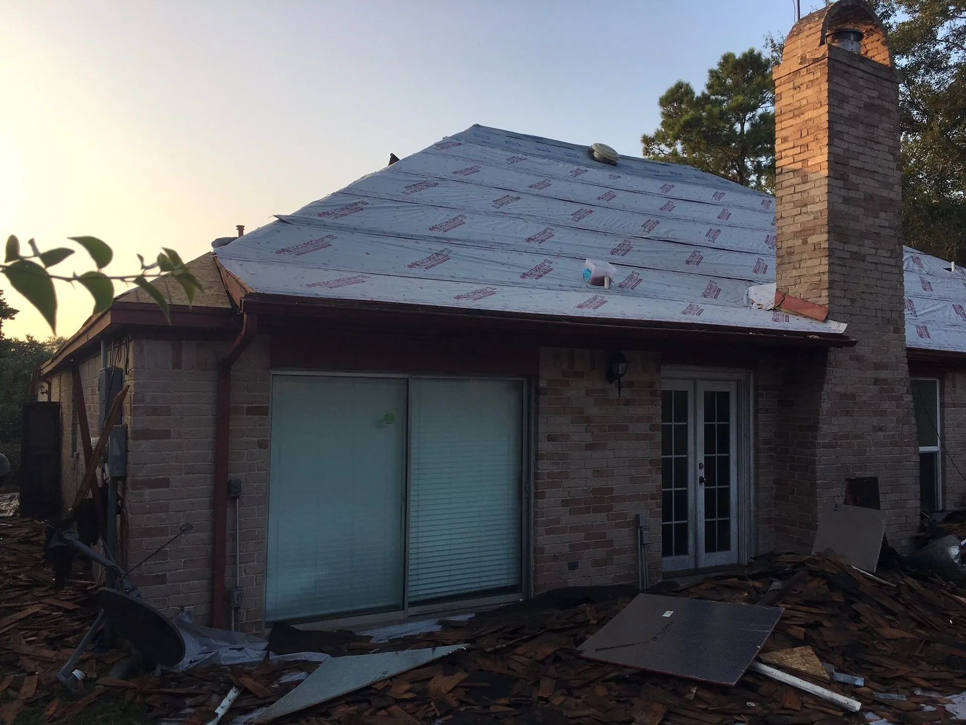 Damaged brick house with collapsed roof and debris in the yard at sunset