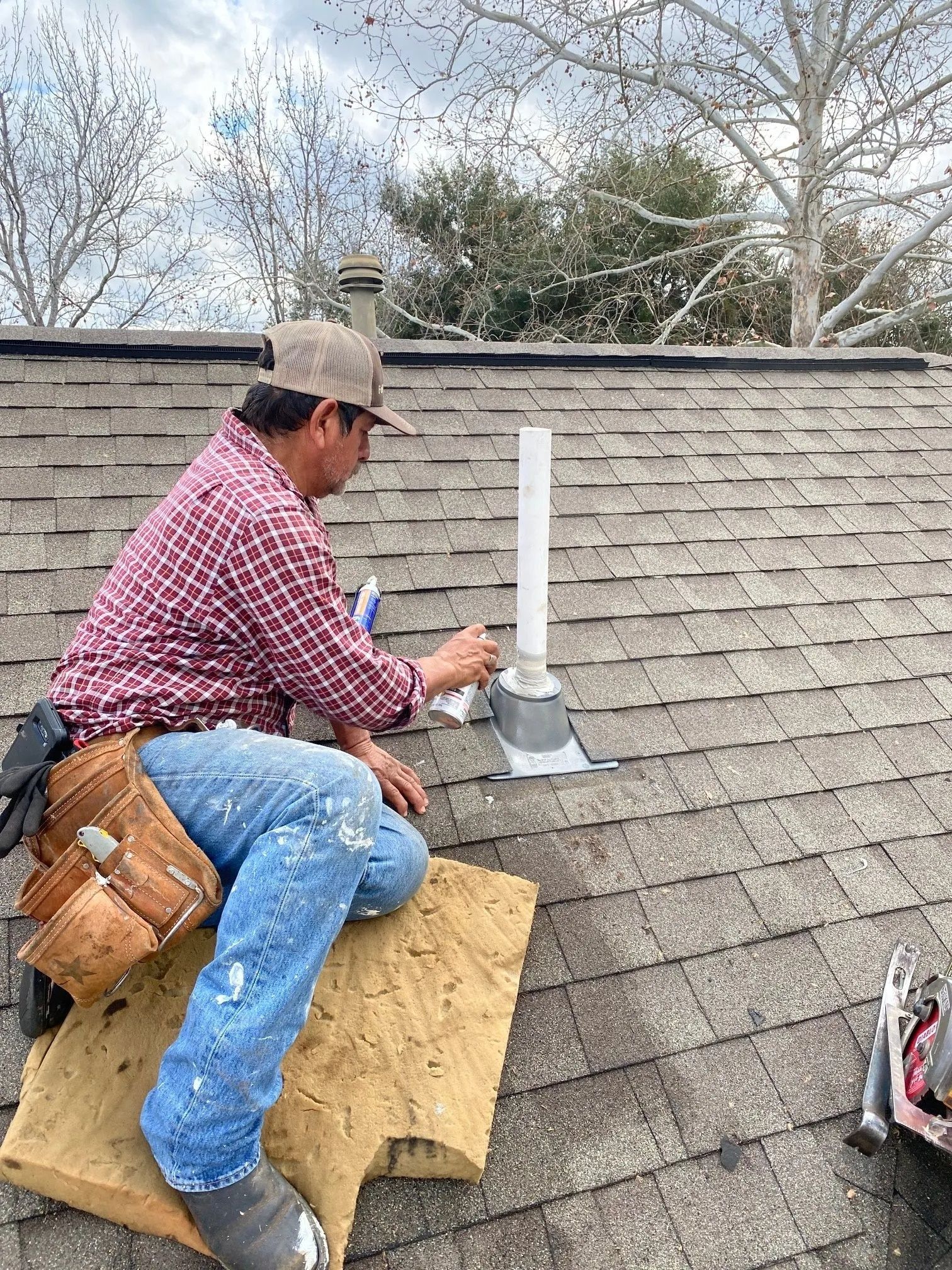 Worker kneels on a roof installing a metal vent pipe in gray shingles.
