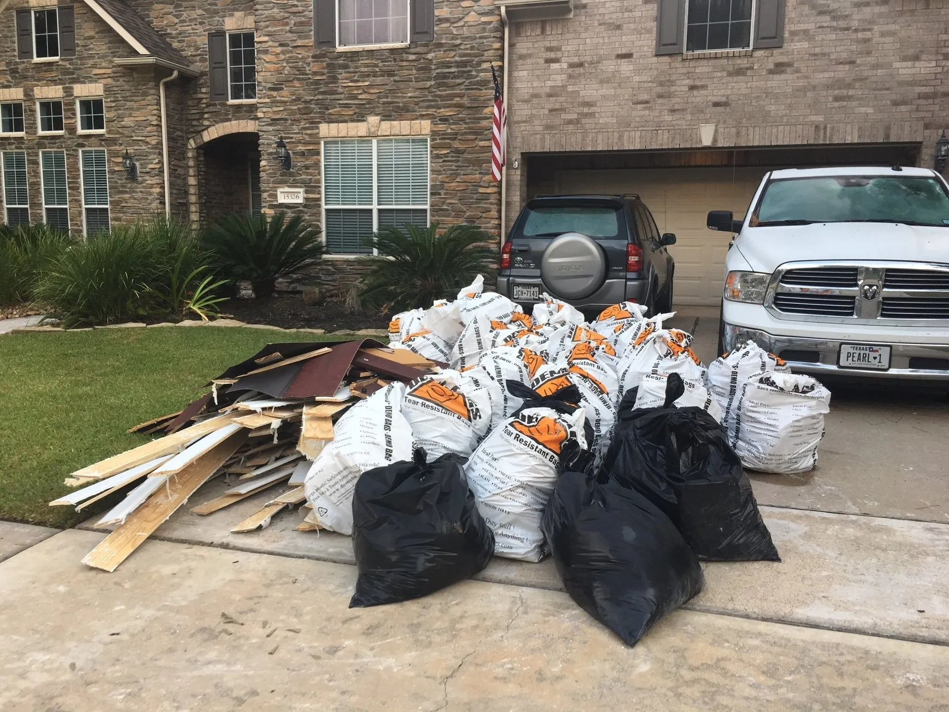 Overflowing trash bags and construction debris piled in a driveway beside parked vehicles outside a brick house