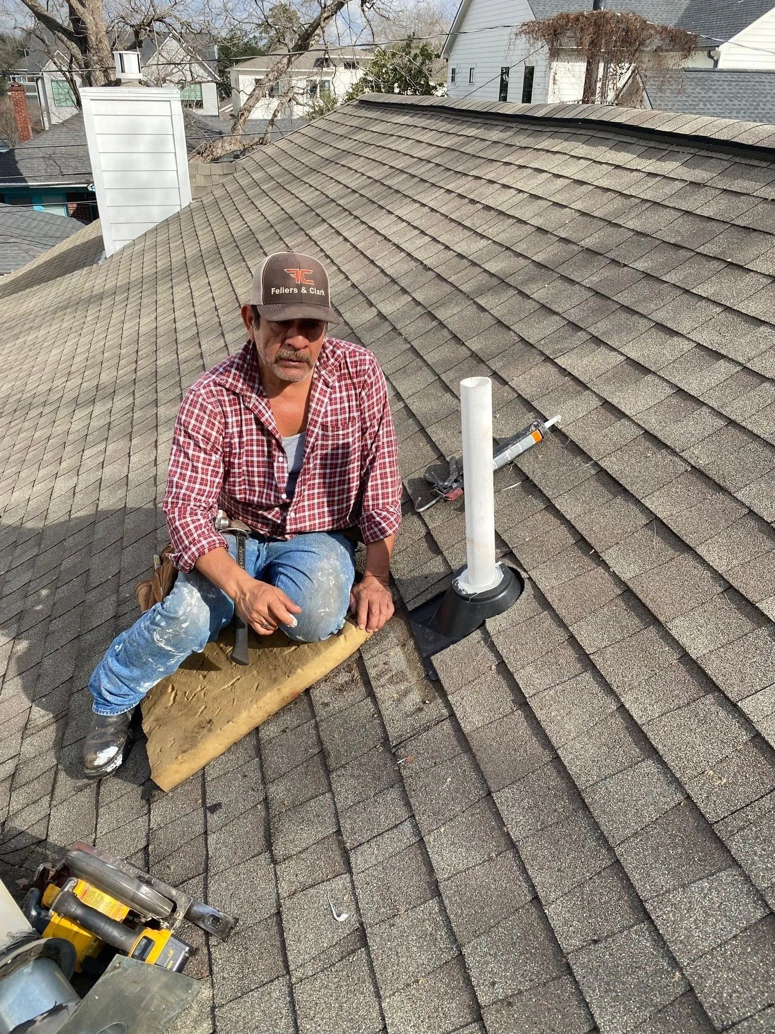 Worker on a shingled roof, sitting beside a vent pipe and holding a wooden plank.
