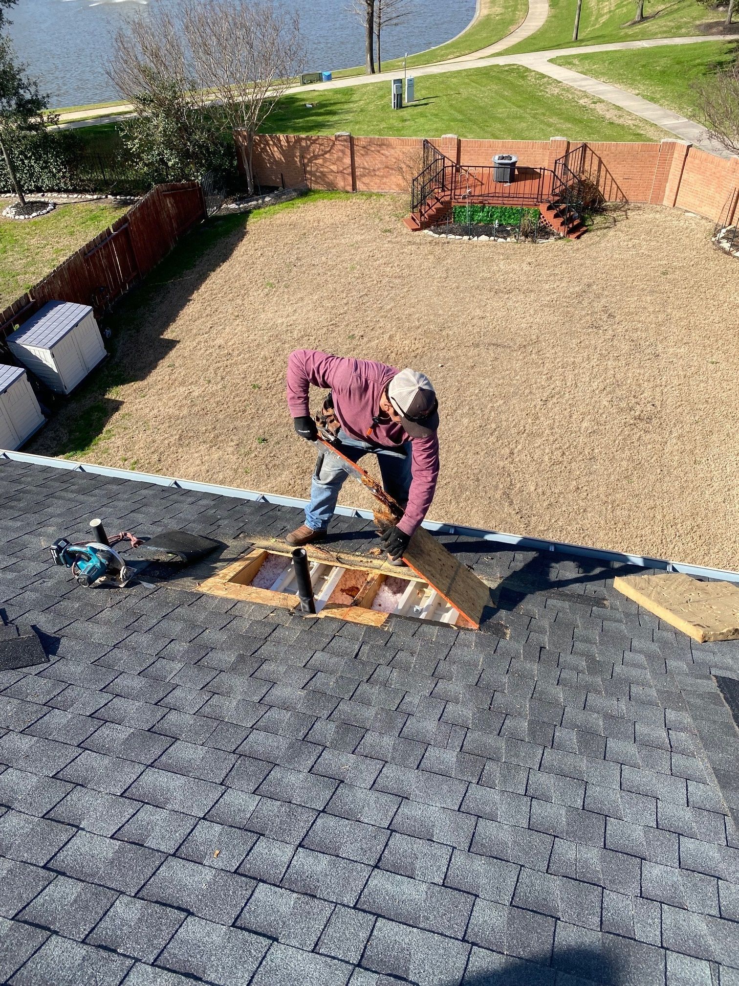 Worker on a roof installing shingles near a gravel backyard and fence