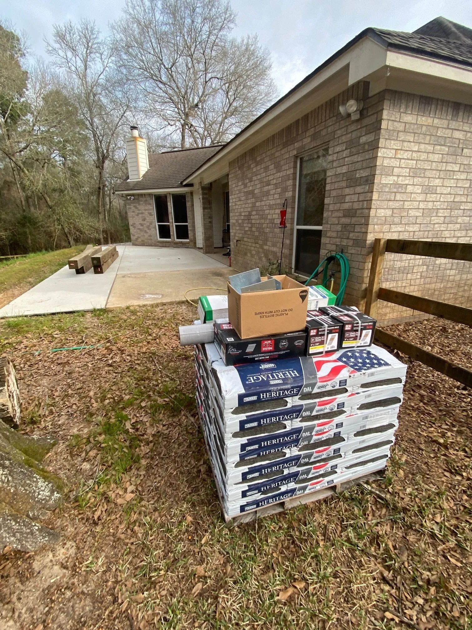 Stacked boxes of flooring on a porch beside a brick house, with tools and a coiled green hose nearby.