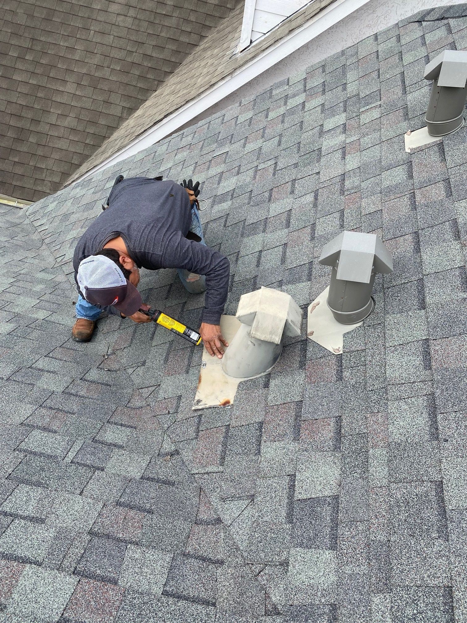 Worker repairing shingles on a roof beside two vents.