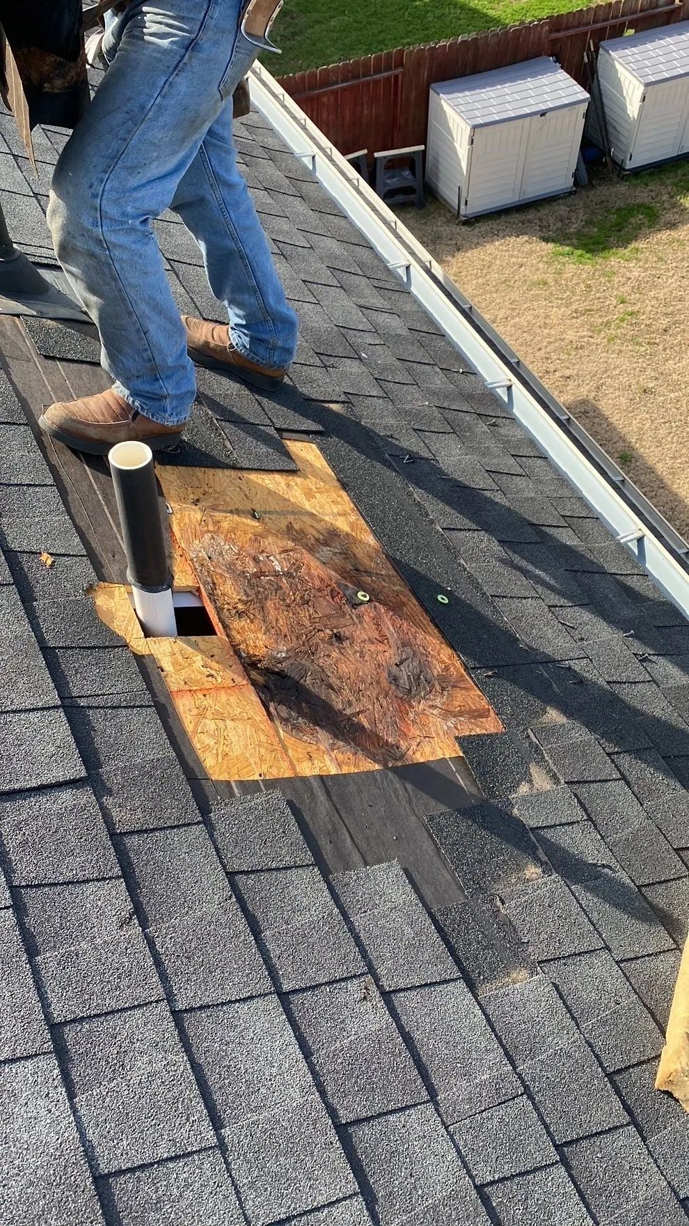 Worker removing damaged shingles from a roof with a pry bar, exposing a rust-stained underlayment.