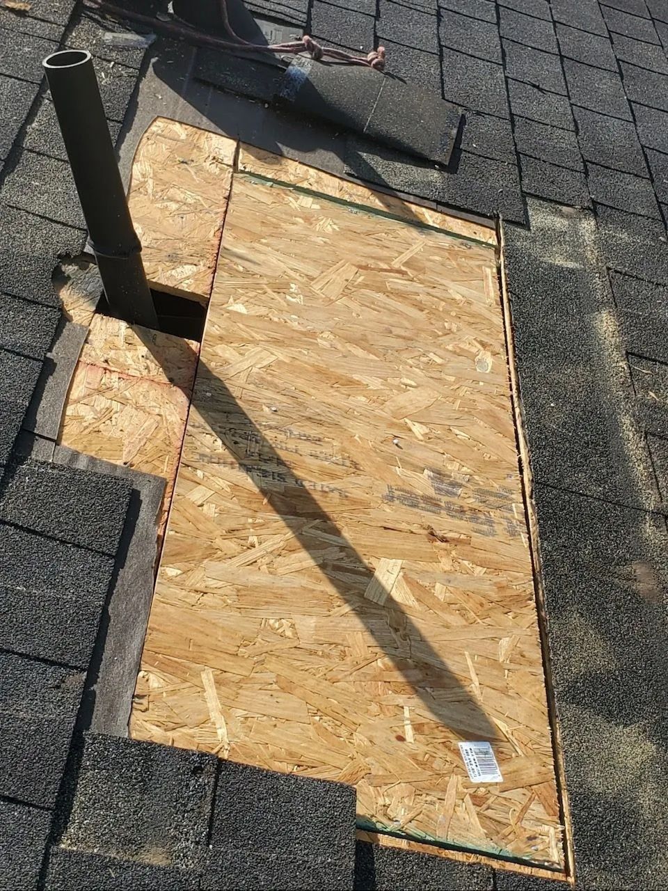 Roof with exposed plywood patch around a vent pipe under dark shingles and sunlight shadows