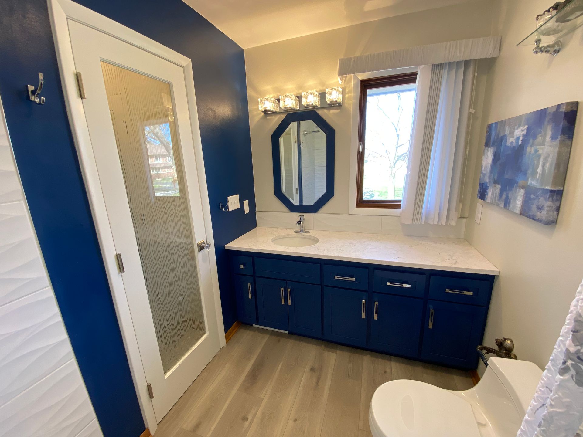 A bathroom with blue cabinets , a toilet , a sink and a mirror.