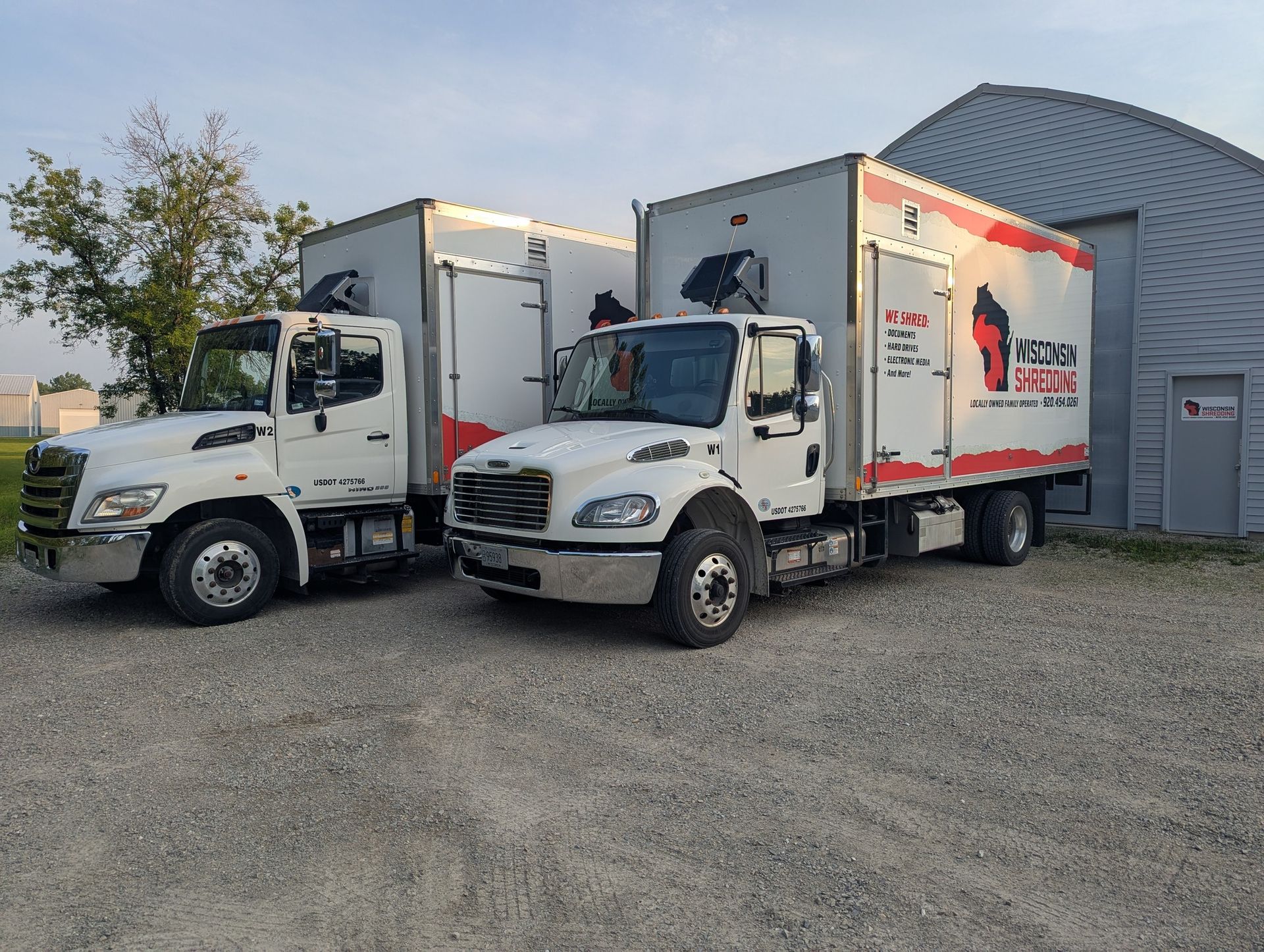 Two white moving trucks parked on gravel, beside a building. One truck has red and black logos.