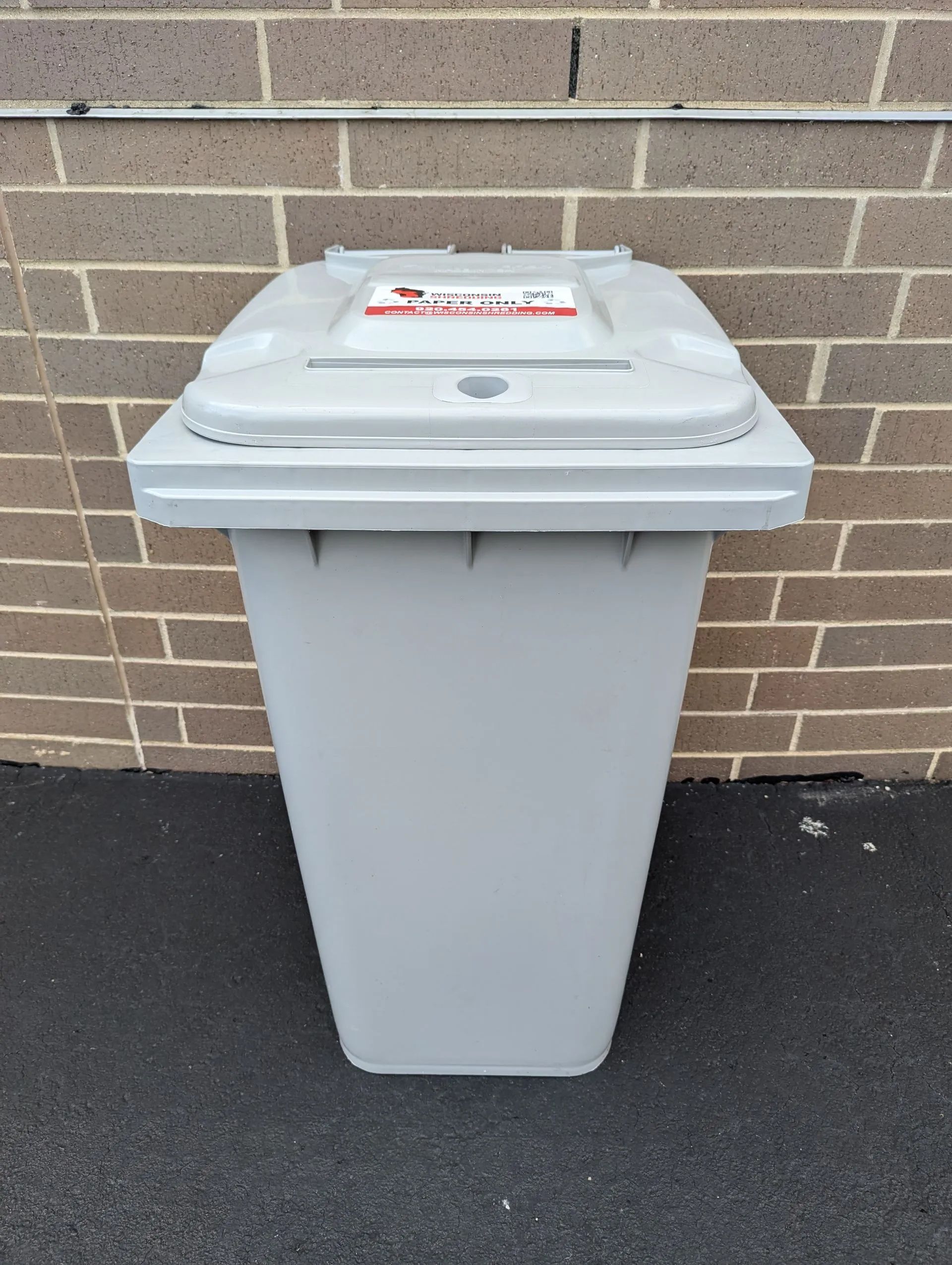 Gray recycling bin against a brick wall.