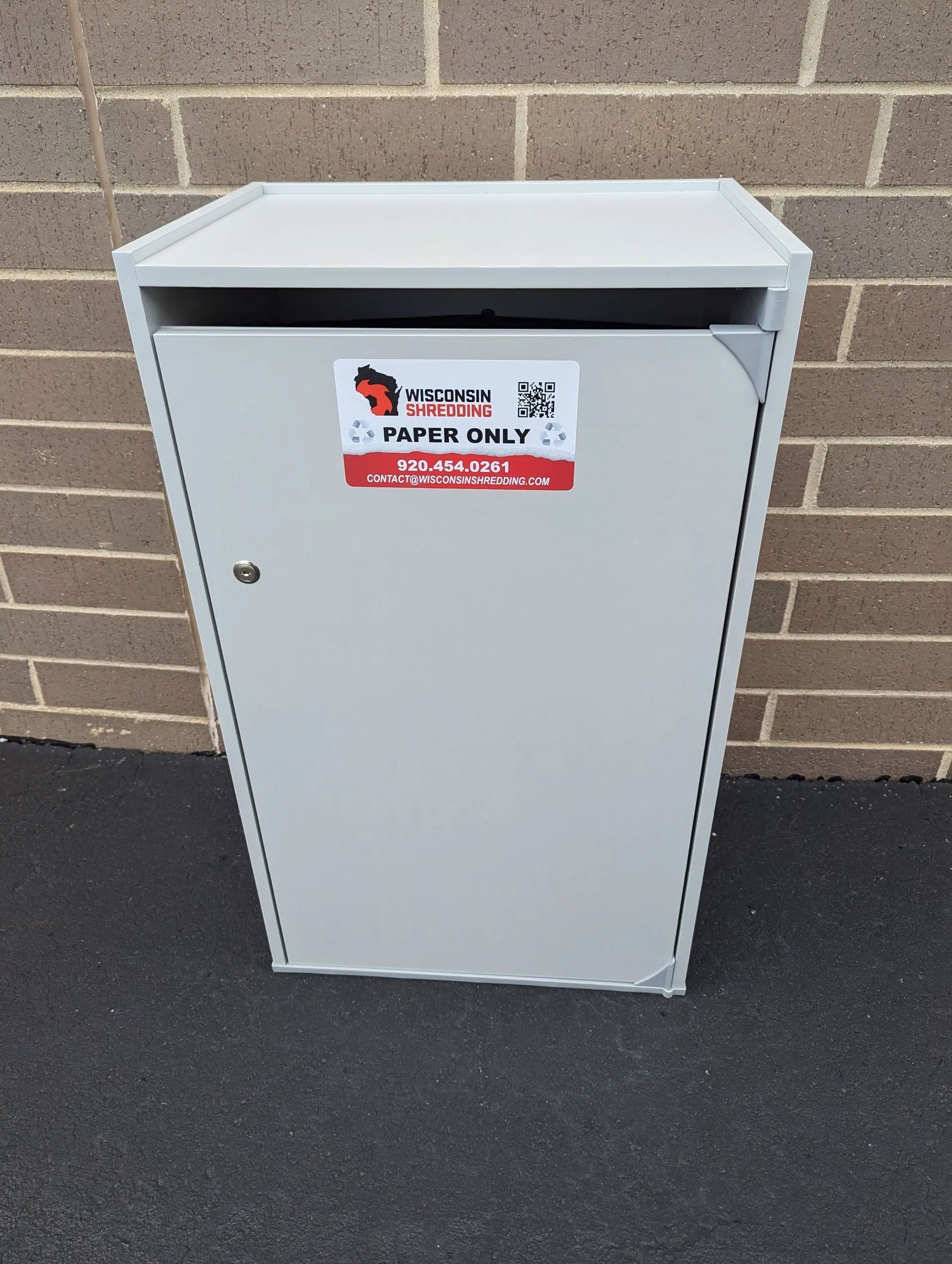 Gray paper recycling bin against a brick wall.  The bin has a 