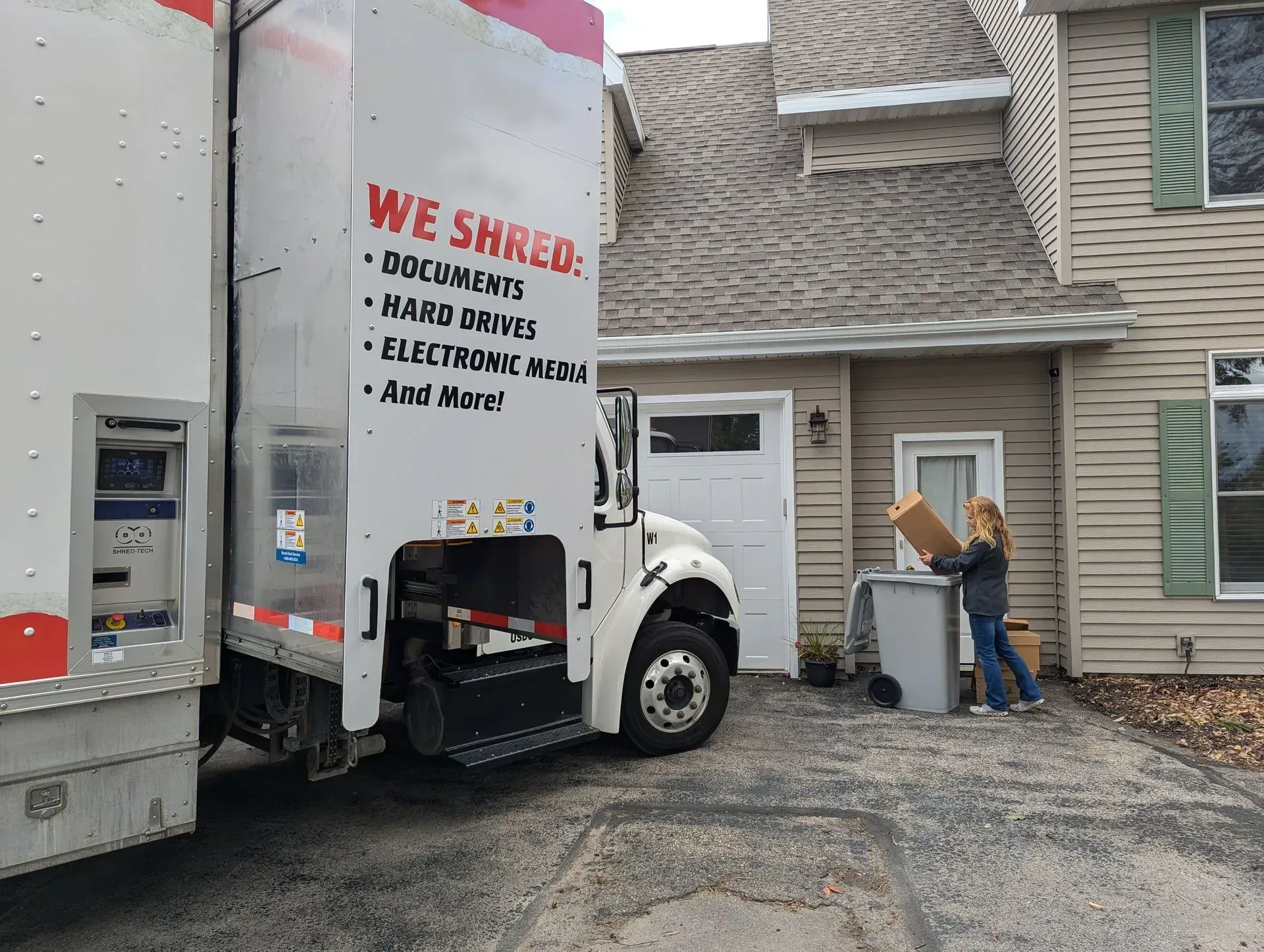 A shredding truck parked in a driveway. A person carries a box toward the truck. A house is in the background.