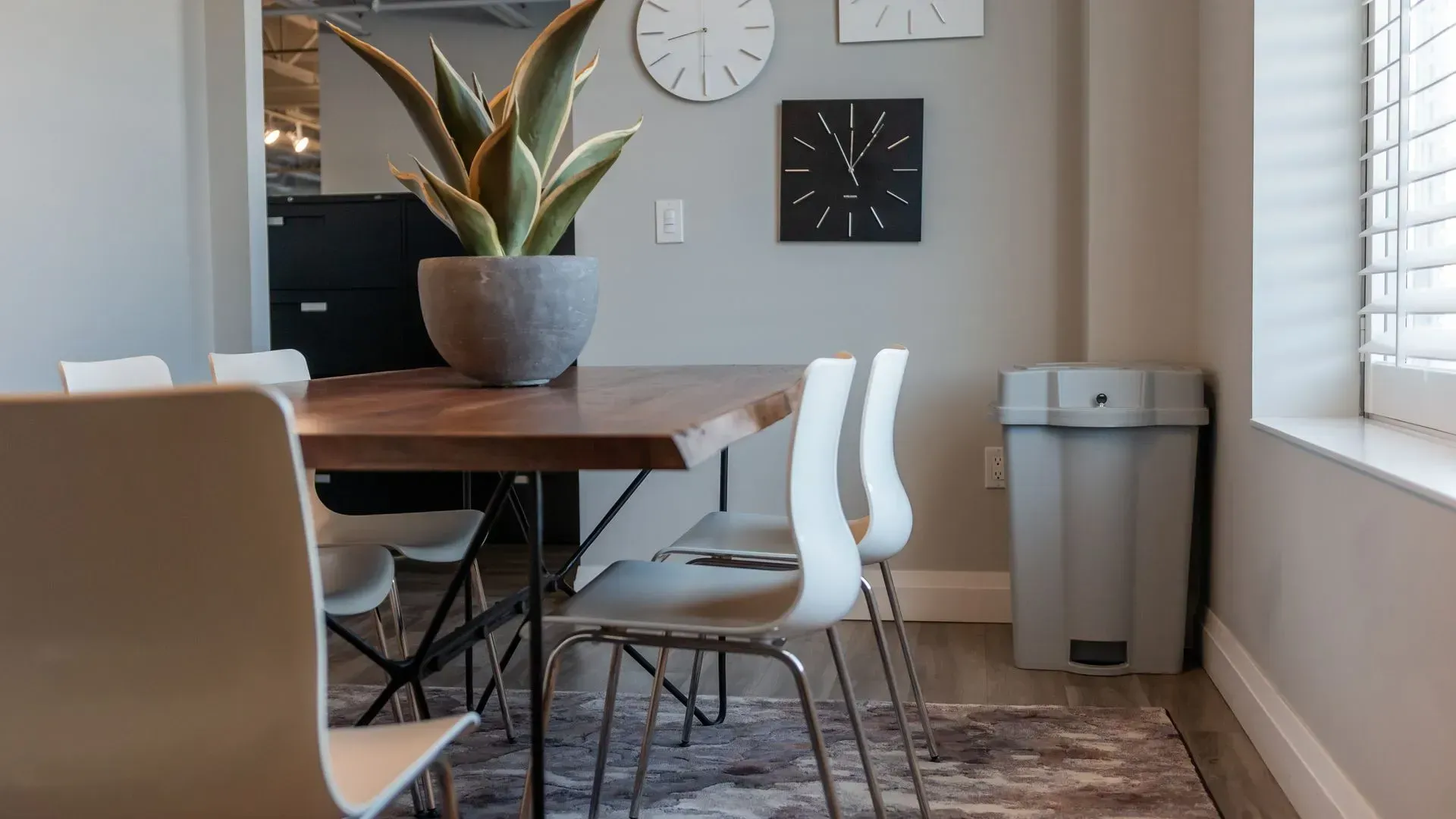 Dining room with a wooden table, white chairs, and a potted plant. Grey walls with clocks and a trash can.