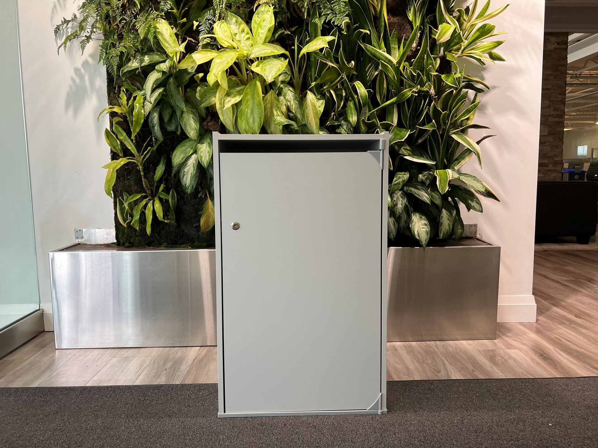Silver mail cabinet in front of a living wall of plants, on a gray mat, and hardwood floor.